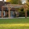 Wooden pergola with a flat slatted roof supported by four timber posts, sheltering a dining table and chairs on a paved patio, set in a landscaped garden with a brick house, white-framed windows, and autumn trees in the background.