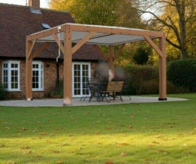 Wooden pergola with a flat slatted roof supported by four timber posts, sheltering a dining table and chairs on a paved patio, set in a landscaped garden with a brick house, white-framed windows, and autumn trees in the background.