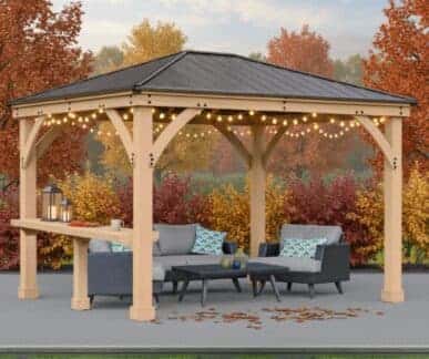 Wooden gazebo with a dark shingled roof and string lights, featuring a gray sectional sofa, dark blue armchair, and black coffee table with patterned throw pillows, set in a landscaped garden with vibrant autumn foliage in red, orange, and yellow.