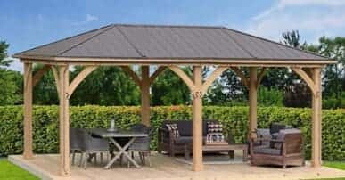Wooden gazebo with metal roof on a timber deck, featuring a round dining table with woven chairs and a cushioned seating area, surrounded by hedges and trees in a landscaped garden.