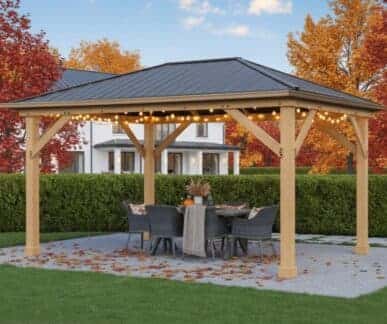 Wooden gazebo with a black metal roof and string lights, featuring a round table with a tablecloth, autumn-themed centerpiece, and six wicker chairs, set on a stone patio surrounded by lawn, hedge, and fall foliage.