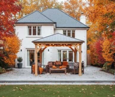 Wooden pergola with string lights and autumn-themed seating area, featuring a sofa and chairs with cushions and throws, set on a stone patio in the landscaped garden of a white house with orange and red trees.