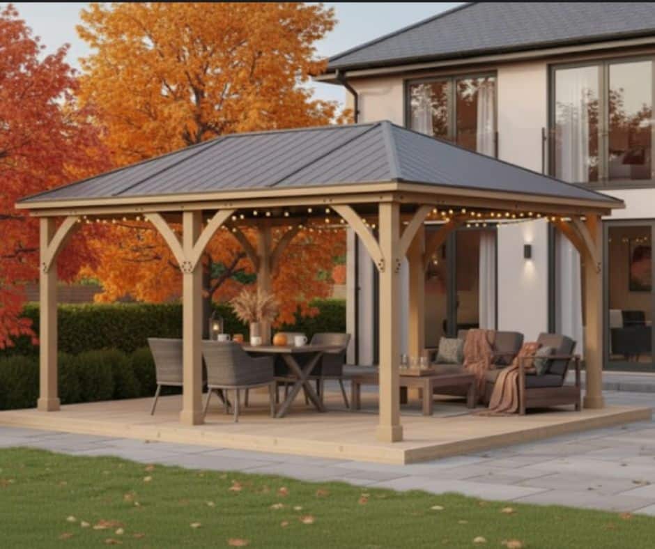 Wooden gazebo with a metal roof and string lights on a paved patio, featuring a dining table, cushioned seating, and autumn foliage, adjacent to a contemporary two-story house.