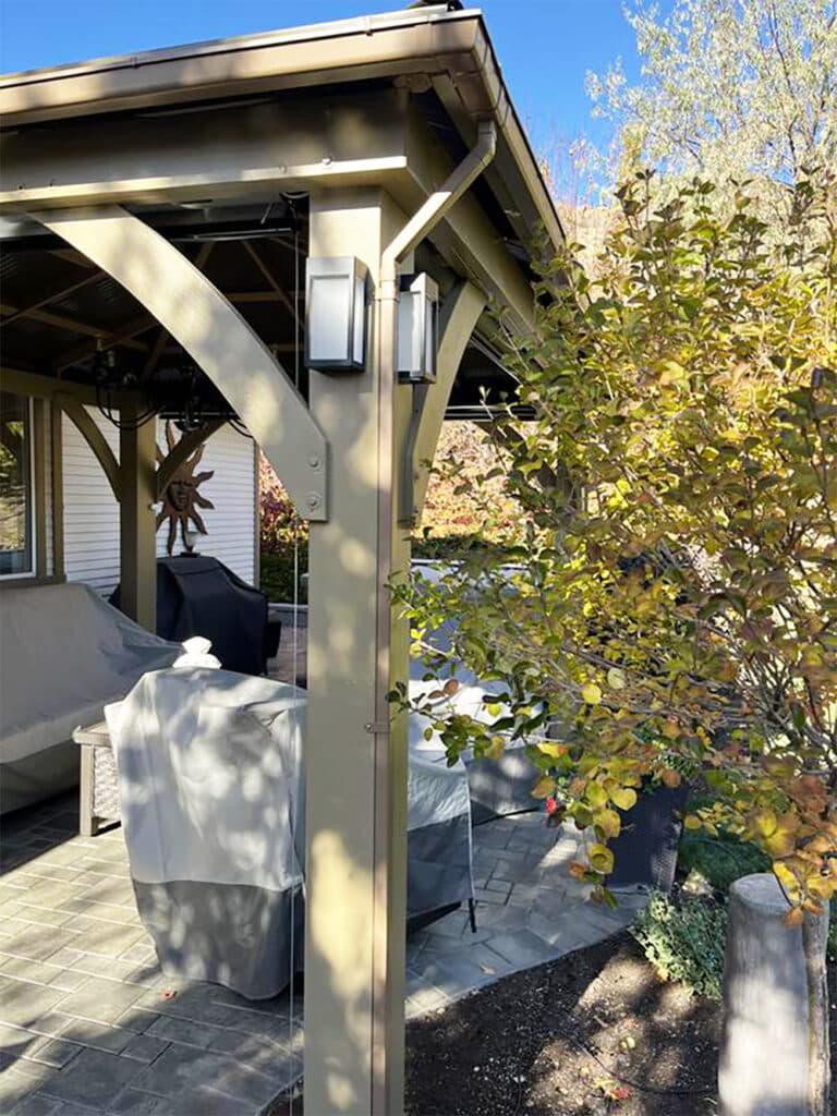 A close-up image of a sturdy, permanent gazebo or pavilion structure built over a brick paver patio on a sunny day. The structure is made of thick, painted metal or composite posts and beams in an olive or tan color. Two vertical outdoor sconce lights with white panes are mounted on the main corner post. Under the gazebo, covered outdoor furniture and a grill, also covered, are visible. A decorative sun wall hanging is mounted on the back wall of a nearby house. To the right, a deciduous shrub or small tree with green and yellowing leaves is prominent. The roof of the gazebo is dark, and the sky is bright blue.
