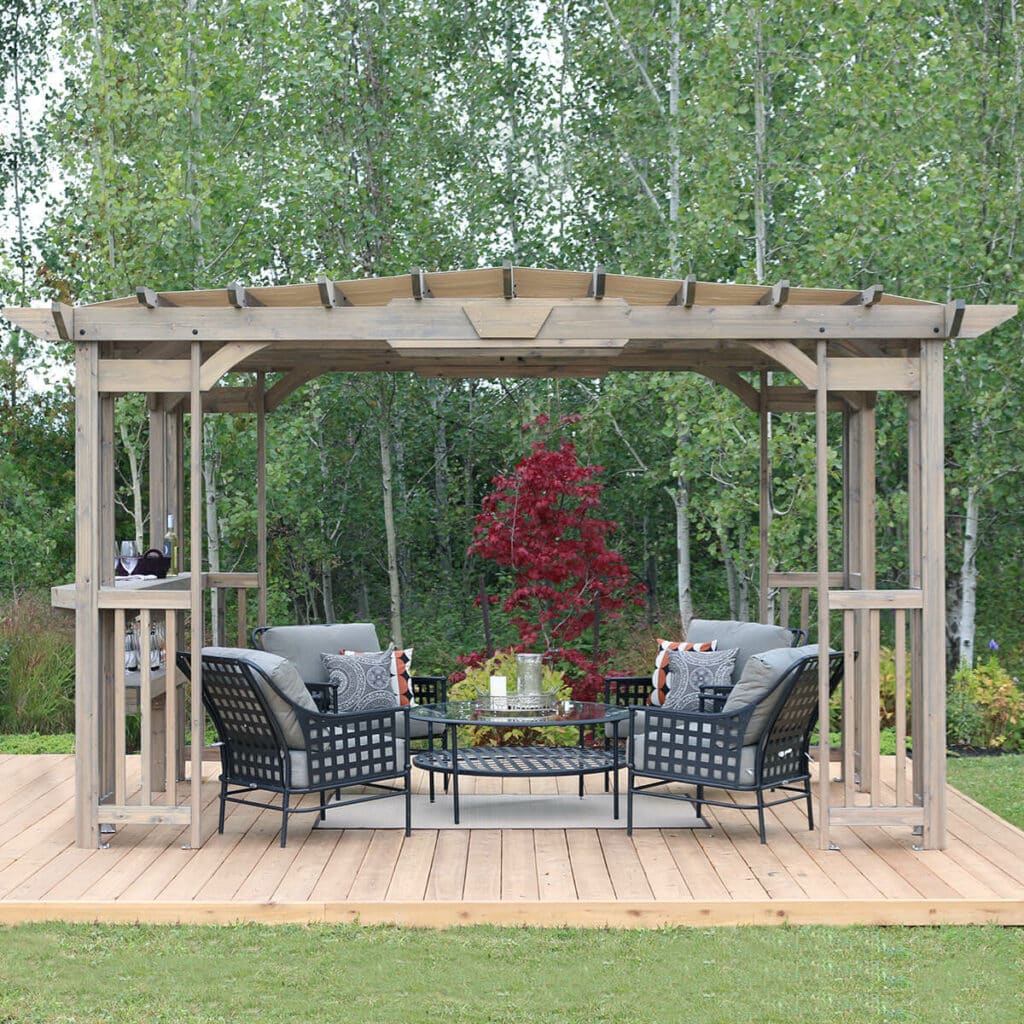 A photo of an outdoor wooden pergola or gazebo structure featuring an outdoor living area. The structure is made of light, weathered wood and has a partially slatted roof. It sits on a wooden deck surrounded by green grass.

Under the pergola, there is a set of dark woven or metal patio furniture with gray cushions and decorative patterned pillows, centered around a glass-topped circular coffee table. On the left side of the structure, there is a built-in wooden bar counter.

In the center background, a striking small tree with vibrant red foliage provides a pop of colour against the backdrop of tall, slender deciduous trees and green undergrowth. The sky is bright but overcast.