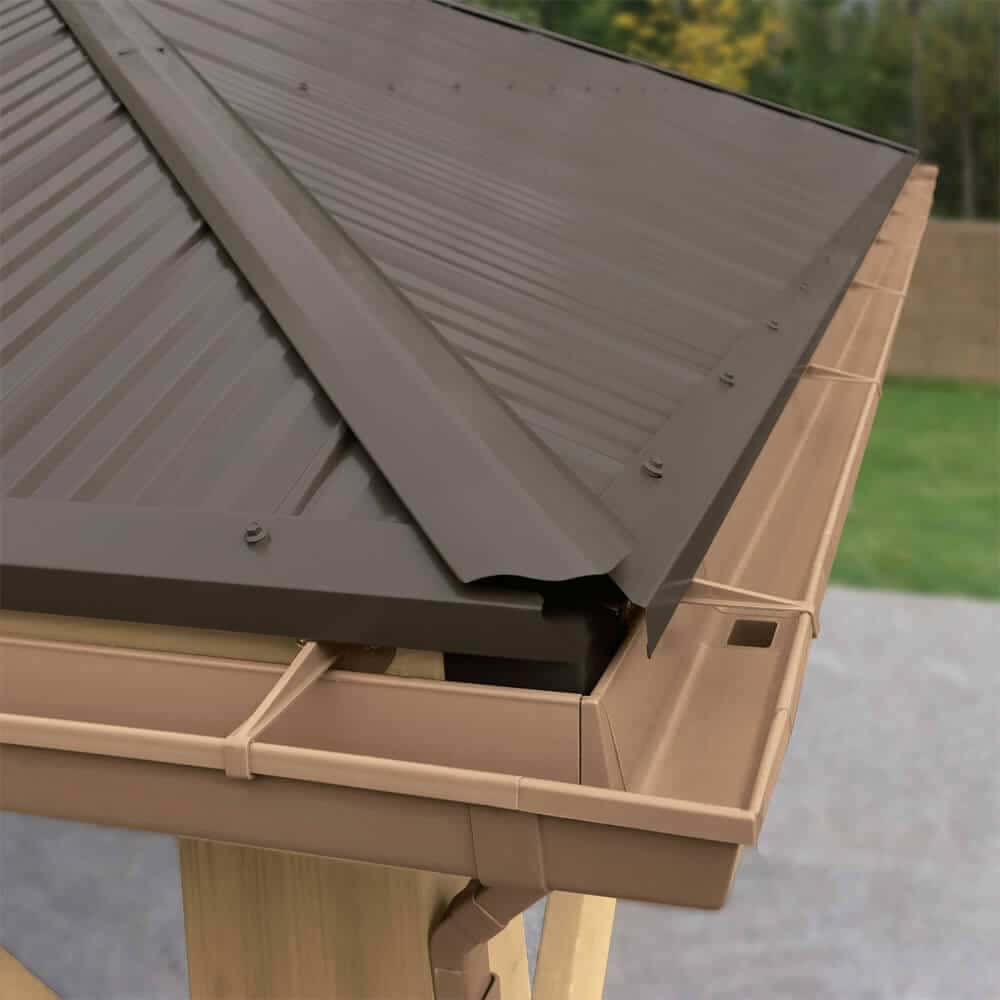 This image is a close-up detail shot of a roof edge and gutter system, likely belonging to an outdoor structure like a gazebo or shed.

Here are the key details:

Roofing Material: The roof is covered with dark brown corrugated metal panels (possibly bronze or dark chocolate brown). The panels are secured with visible fasteners (screws). The structure has a pitched roof, with a prominent ridge cap visible at the corner.

Gutter System: A complete light brown/tan gutter system (likely vinyl or plastic) is installed along the edge of the roof.

It includes a corner piece and appears to have a downspout connection visible near the corner.

The gutter color provides a warm contrast to the dark roof.

Support Structure: A portion of the underlying wooden frame or trim is visible supporting the roof and gutter.

Background: The roof extends over a smooth light grey patio or paving area, with a hint of green lawn and a blurred background of trees/foliage.

The image showcases how a finished, weather-tight roof and water management system is installed on an outdoor structure.