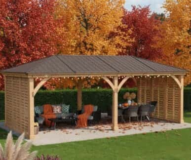Wooden gazebo with a dark brown metal roof and decorative arches, featuring a lounge area with a sofa and coffee table, and a dining setup with chairs, set on a paved patio surrounded by autumn foliage and a green hedge.