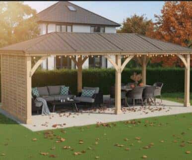 Large wooden gazebo with a metal roof and string lights, featuring a sectional sofa, coffee table, and dining setup on a paved patio, surrounded by green grass and autumn trees near a modern two-story house.