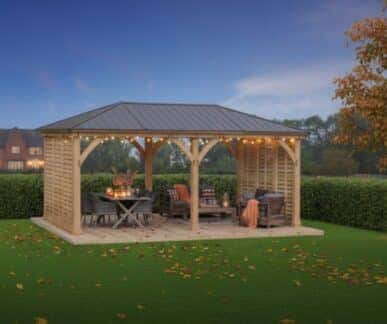 Wooden gazebo with a dark pitched roof and lattice-style side panels, featuring cushioned seating, a table with autumn decorations, and string lights, set in a grassy yard with fall foliage and houses in the background.