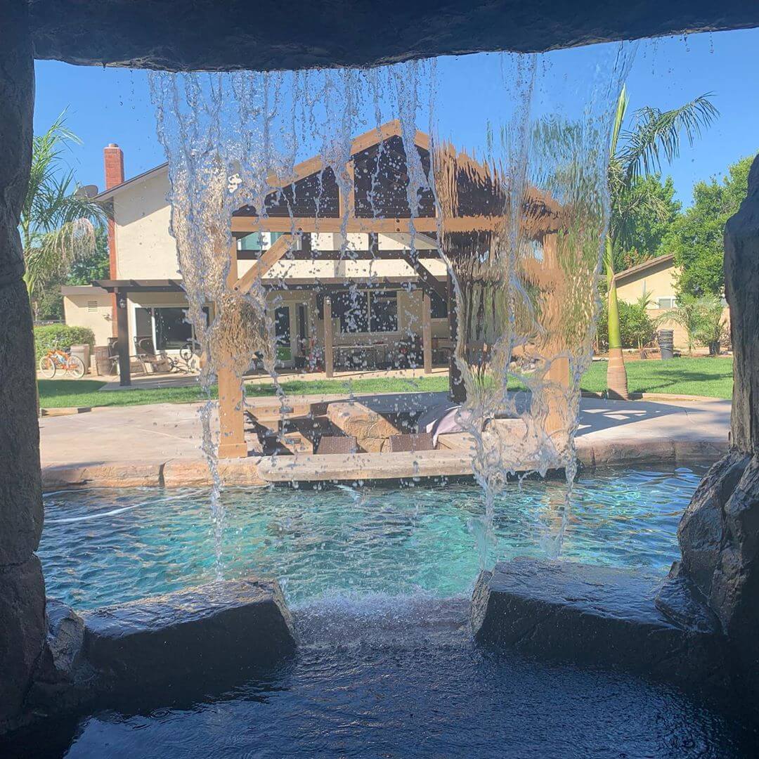 View from behind waterfall feature in swimming pool looking out to backyard patio, pergola, and house.