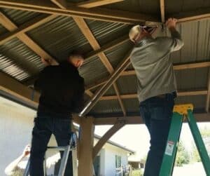 Two people installing corrugated metal roof panels on wooden gazebo frame using ladders and power tools
