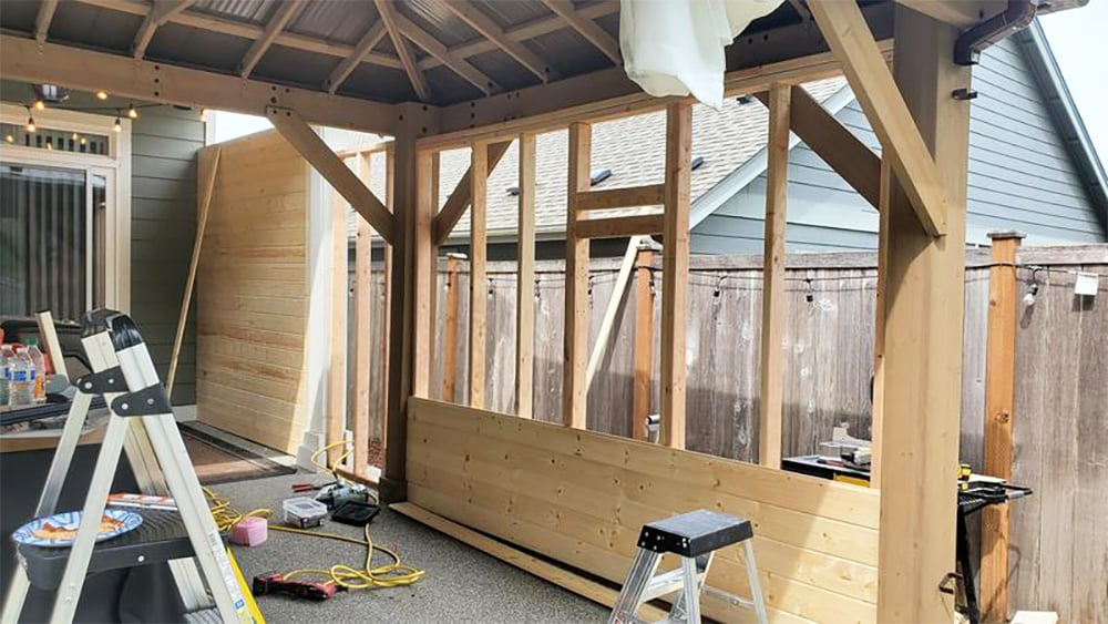 This image captures an in-progress construction project—specifically, enclosing an open wooden outdoor structure (like a pergola or pavilion) to create a more sheltered space.

Here is a detailed breakdown of the scene:

Main Structure: A sturdy wooden frame (posts and beams) is visible, with a translucent or corrugated roof covering overhead, suggesting it was originally a patio cover or open gazebo.

Construction Work:

Walls: New vertical wooden studs have been framed within the existing posts to create a wall. A partial wall of tongue-and-groove horizontal cedar or pine boards is being installed along the bottom. A finished board wall is already in place on the left side of the structure.

Tools & Equipment: The ground is scattered with construction materials and tools, including:

Extension cords (yellow).

Various power tools (drills, saws).

A couple of step ladders or step stools.

A collection of debris and possibly some snacks (a plate with food remains on the small ladder to the left).

Background & Setting:

The structure is attached to or immediately next to a house with light green or sage siding and a white door/window frame.

Beyond the structure is a wooden privacy fence and the roofline of a neighboring house.

Atmosphere: The image conveys a sense of active DIY work and transformation of an outdoor space into a more private, functional room.