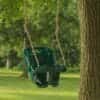 Green plastic baby and toddler swing with a high back and front support, suspended by ropes from a tree branch over a grassy lawn with trees in the background.