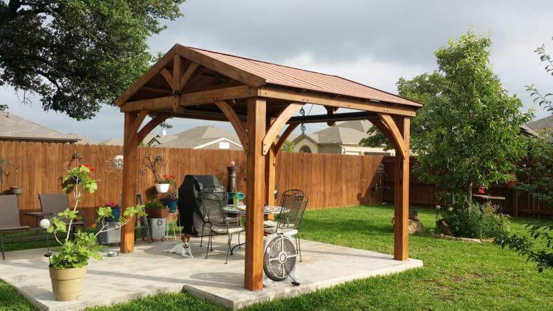 A beautiful wooden gazebo or pergola with a solid, metal (or possibly shingled) roof, creating a great outdoor living space!
A freestanding wooden structure, likely cedar or treated pine, with robust posts and arched braces at the top.
A pitched roof with a brown/bronze colour, offering full protection from sun and rain.

Patio: The structure is set on a concrete or paved patio area.
It serves as an outdoor dining/grilling area, containing a BBQ grill, a table, and chairs.
 It's located in a well-maintained backyard surrounded by a wooden privacy fence and green lawn.

It seems like a perfect spot for entertaining or relaxing outdoors!