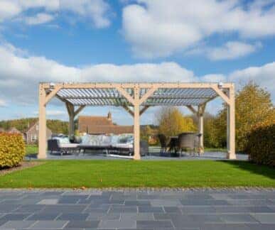 Large wooden pergola with a slatted roof, featuring cushioned sofas and a dining table with chairs, set on a dark gray tiled patio surrounded by green grass and trimmed bushes in a suburban garden.