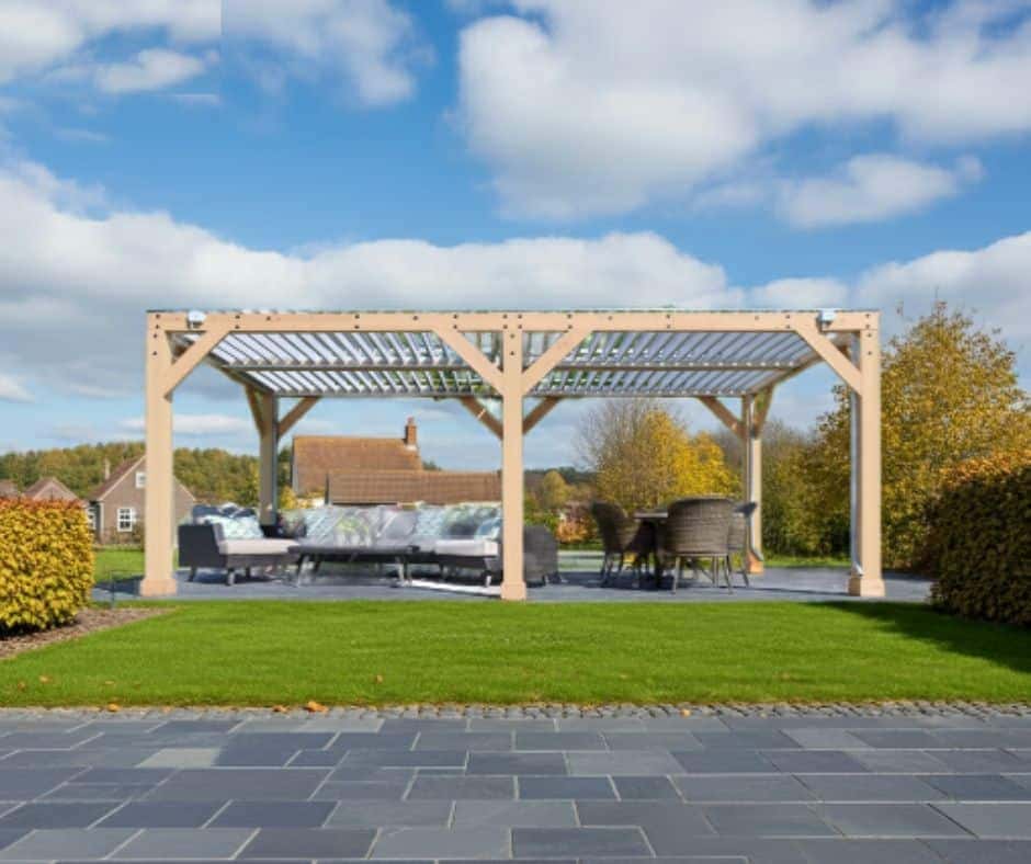 Large wooden pergola with a slatted roof, featuring cushioned sofas and a dining table with chairs, set on a dark gray tiled patio surrounded by green grass and trimmed bushes in a suburban garden.