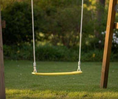 Yellow plastic swing suspended by white ropes from a wooden frame, set on a grassy lawn with trees and foliage in the background.