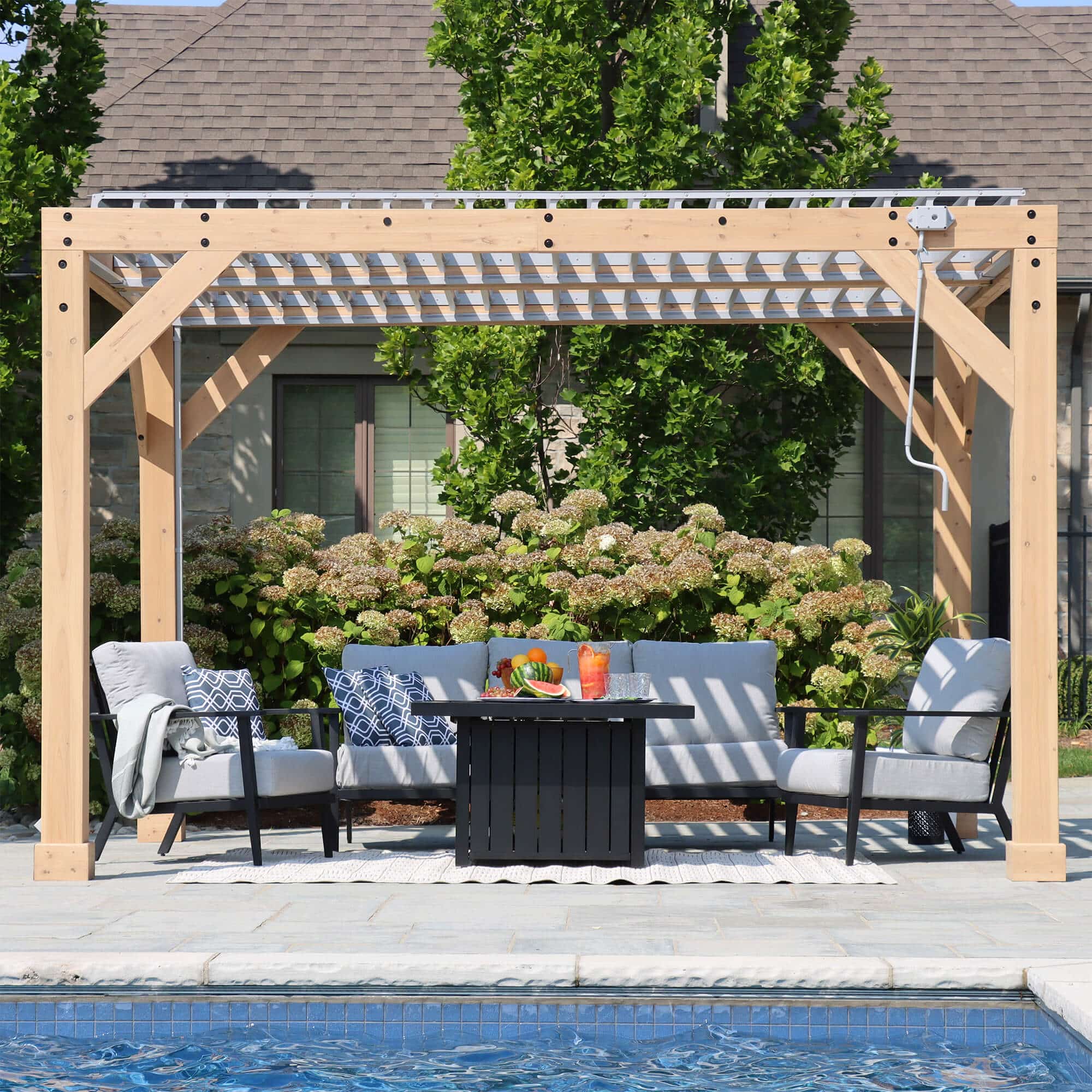 Wooden pergola with slatted roof over cushioned armchairs and fire pit table, set beside a swimming pool with lush garden and hydrangeas in the background.