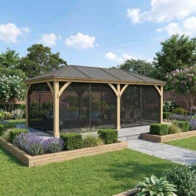 A large wooden gazebo with a dark metal roof and screened sides sits in a landscaped garden with stone pathways, raised planters, trimmed grass, and surrounding trees and flowering shrubs.