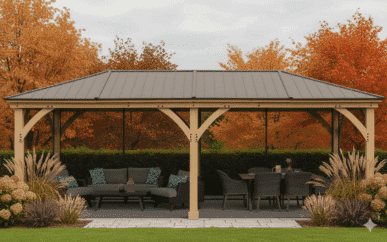 Large wooden gazebo with a metal roof, featuring a sectional sofa with patterned cushions and a dining table with six chairs, set on a stone patio surrounded by ornamental grasses, trimmed hedges, and autumn trees with red and orange foliage.