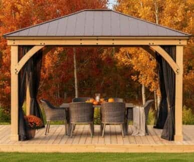 Wooden gazebo with a metal aluminium roof and sheer black mesh curtains, set on a deck with a dining table decorated with pumpkins and a candle, surrounded by wicker chairs and autumn foliage.