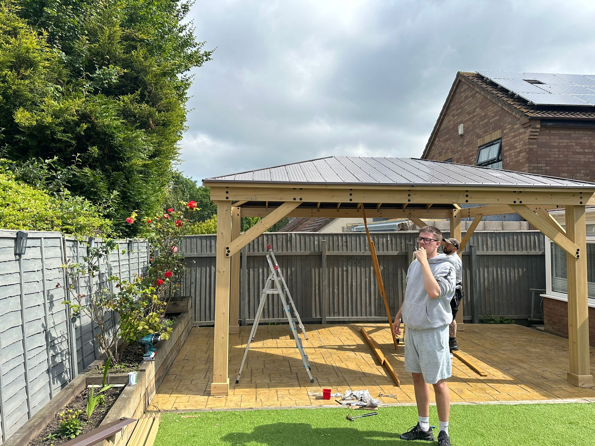 Backyard gazebo construction scene with metal roof, ladder, paint supplies, and two people working on paved patio