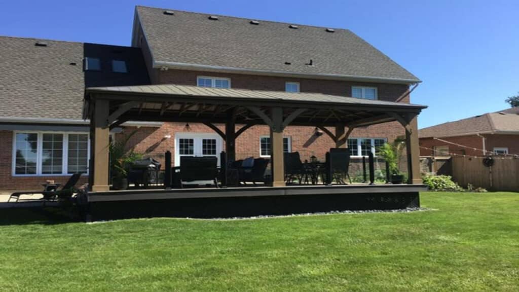 This image features a stylish, freestanding outdoor gazebo built on a raised deck, situated directly behind a large residential home.

Here is a detailed description:

Gazebo/Pavilion:

The structure is a large, open-sided gazebo with a dark brown/bronze metal roof.

It has a sturdy wooden or composite frame in a darker brown/tan color.

It shelters a complete outdoor living space, including dark-colored outdoor seating (sofas, armchairs) and a dining table set beneath it.

Potted plants are visible flanking the structure, adding greenery.

Deck:

The gazebo is built on a raised deck, which appears to be constructed from dark, possibly composite, decking material.

The perimeter of the deck features a dark railing with clear or black vertical balusters and a dark top rail.

The exposed lower edge (skirting) of the deck is also dark, creating a clean, defined look against the lawn.

A ramp or steps (partially visible on the left) provides access from the yard.

House and Background:

The structure is positioned behind a large, two-story brick house with several windows and white French doors leading out to the deck.

The house has a dark brown shingle roof.

The surrounding area includes a well-maintained green lawn in the foreground and a lower brown fence dividing the property from a neighboring house on the right.

The overall scene depicts a well-designed, integrated outdoor entertaining area that extends the living space of the home into the backyard.