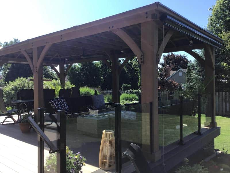 This image depicts a cosy and modern outdoor living space featuring a large wooden gazebo built on a raised deck, overlooking a backyard.

Here is a detailed description:

Gazebo/Pavilion: The structure has a robust frame made of dark-stained or naturally dark wood, featuring thick posts and diagonal braces (like the previous images). The roof appears to be flat or low-pitched and finished in a dark color.

Outdoor Living Area: Under the gazebo, there is a comfortable seating area with dark-coloured outdoor furniture, including chairs and a sofa with patterned cushions.

Deck and Railing:

The gazebo is built on a raised deck, which appears to be a dark composite or stained wood, providing a unified, contemporary look.

The most distinct feature is the modern railing system. It consists of tempered glass panels held by dark (black) metal posts and handrails. This glass railing provides safety while maximising the view of the surrounding yard.

The deck is accessed via a small flight of stairs visible in the foreground, also featuring the glass railing.

Decor and Ambiance: A light-coloured, tall woven rattan lantern or decorative piece sits on the deck, adding a natural and ambient touch to the dark colour scheme. Potted plants are also visible, helping to blend the structure with the landscape.

Background: The structure faces a lush, green backyard with mature trees and landscaping, and the roofline of a neighbouring house is visible in the distance. The bright sun suggests a clear, pleasant day.

The overall scene showcases a private, stylish, and well-designed outdoor retreat ideal for relaxation and entertaining.