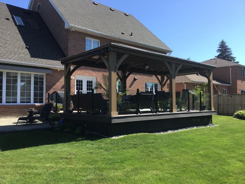 Timber pergola with gabled roof on raised deck beside brick house, featuring outdoor furniture and glass railings