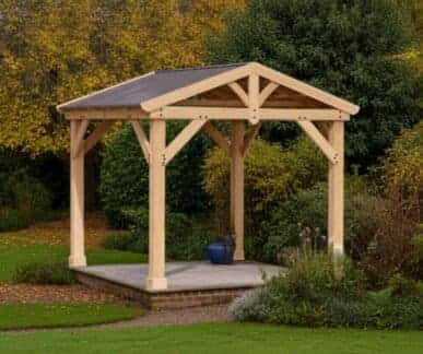 Wooden gazebo with a dark pitched roof supported by six timber posts, set on a raised stone platform bordered by bricks, surrounded by autumn trees, shrubs, and flowering plants in a landscaped garden.
