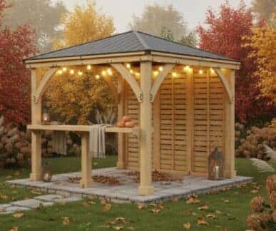 Wooden gazebo with a dark shingled roof and string lights, featuring a counter with pumpkins, set on a stone patio surrounded by autumn foliage, ornamental grasses, and lanterns.