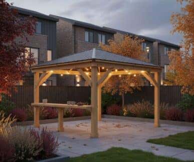 Wooden gazebo with a pitched metal roof and string lights, featuring a long table set with cups and plates, placed on a paved patio surrounded by landscaped garden beds and autumn trees, with brick residential buildings in the background.