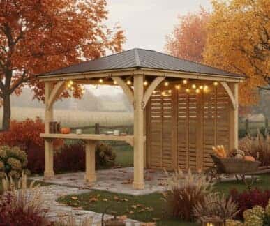 Wooden gazebo with a dark metal roof and string lights, featuring a long bar-height counter with pumpkins and kitchenware, surrounded by autumn trees, ornamental grasses, and a stone path in a landscaped garden.