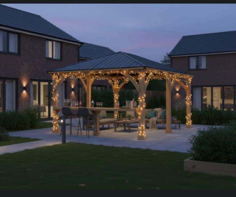 Wooden gazebo with a dark metal roof and string lights, featuring a long dining table with benches and chairs, set with glasses and bottles on a stone patio, surrounded by lawns and brick houses at dusk.