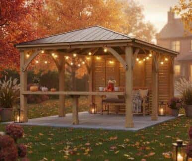 Wooden gazebo with a pitched metal roof and string lights, featuring a cushioned bench, table with beverage dispenser, and autumn decorations including pumpkins and lanterns, set on a stone patio with fall foliage in the background.