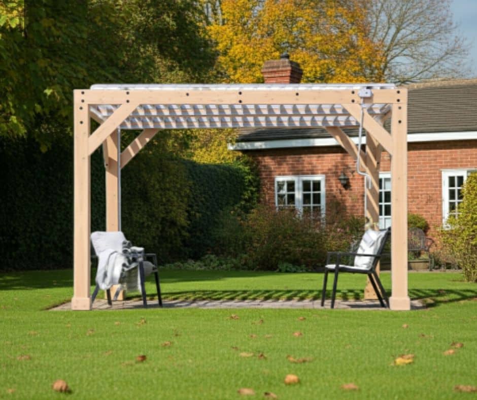 Wooden pergola with a slatted roof casting patterned shadows, featuring two black chairs with light-colored blankets, set on a lawn in a landscaped garden with a red brick house and autumn trees in the background.