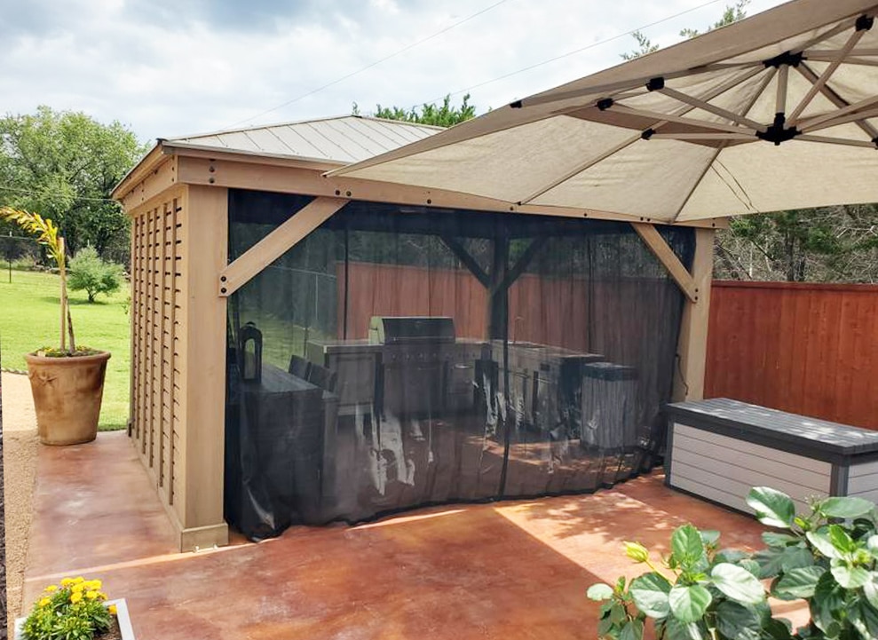 Outdoor kitchen under timber-framed gazebo with metal roof, mesh curtains, stainless appliances, and canopy shade