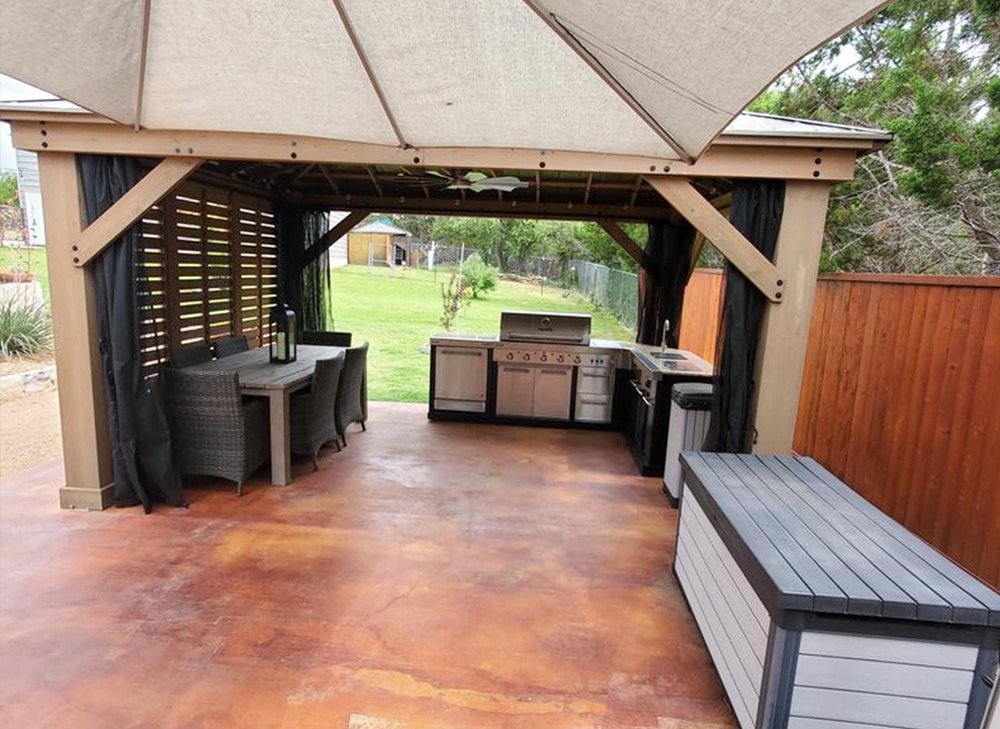 This image captures a dedicated, fully-equipped outdoor kitchen and dining area housed within a large pavilion structure.

Here is a detailed description:

Structure (Pavilion/Gazebo):

It has a sturdy, light-brown/tan composite or wooden frame with thick posts and diagonal bracing.

The roof is partially visible and appears to be covered by a large, light-colored fabric or canvas awning (similar to a cantilever umbrella or sun shade) extending beyond the main frame.

The sides are equipped with dark, black fabric curtains that can be drawn for privacy or shade.

The left side has decorative horizontal wooden slats for partial enclosure.

Outdoor Kitchen:

A complete built-in kitchen unit runs along the back wall.

It features stainless steel appliances, including a large grill/BBQ in the center, and a sink on the right.

The cabinets and counter surrounds are a light color (possibly white or gray composite/metal).

A ceiling fan is visible in the center of the roof structure above the kitchen area.

Dining Area:

On the left side, there is an outdoor dining set with a dark, rectangular table and several woven (possibly rattan or wicker) chairs. A decorative lantern sits on the table.

Flooring and Storage:

The floor is a spacious concrete or paved patio with a striking red/brown, rust-colored stain or finish.

In the foreground on the right, there is a large, two-toned gray and white outdoor storage bench/box (similar to the one in the previous image).

Background:

The pavilion opens up to a grassy yard bordered by a wooden fence (brown on the right) and some trees in the distance.

The overall scene depicts a highly functional, well-designed, and comfortable outdoor living space.