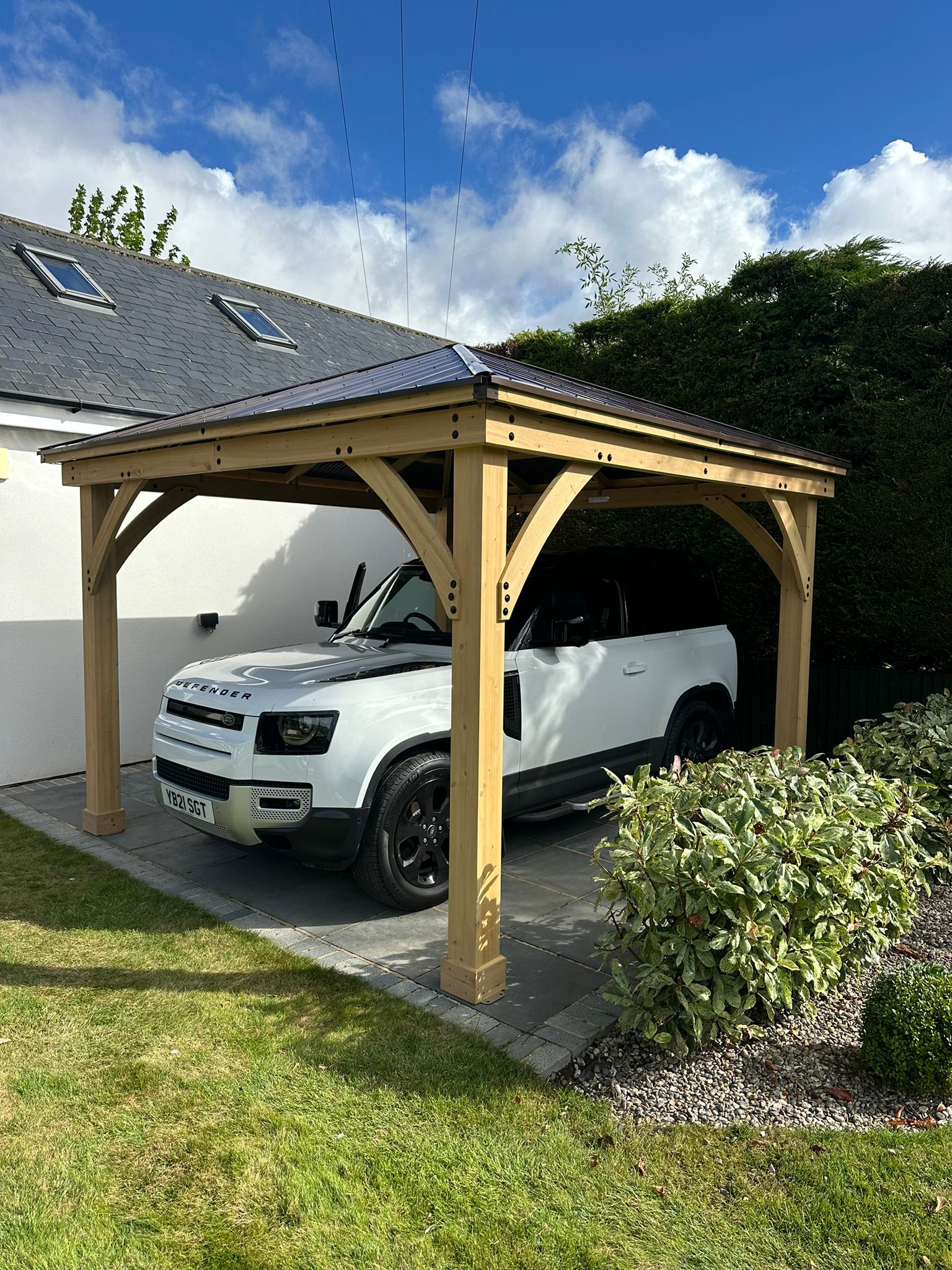 Timber carport with pitched shingle roof sheltering white Land Rover Defender beside white house