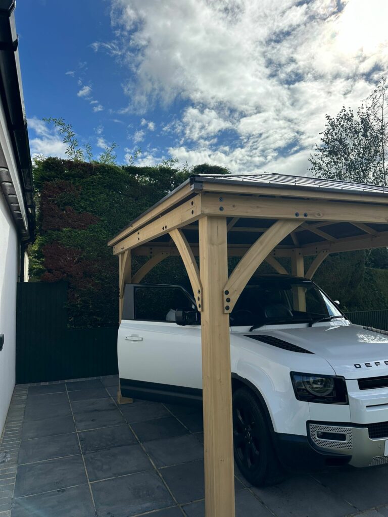 This image features a white modern Land Rover Defender 90 (two-door model) parked under a newly built, attractive wooden Yardistry cedar wood gazebo structure.

Key Details:
The Carport: The structure is made of light-coloured, pre stained premium cedar wood, with robust support posts and curved braces. The roof appears to be dark brown, aluminium easy care, with some sunlight reflecting off it. It is situated over a paved driveway area.

The Vehicle: A white Land Rover Defender with a black roof and black wheels is parked neatly beneath the canopy. The car appears washed clean and polished.  

Setting and Landscaping:

The structure is adjacent to a white house with a dark, tiled roof that features multiple skylights.

The area is well-landscaped, with a stretch of green, manicured lawn in the foreground and a clipped dark green hedge forming a backdrop behind the structure.

In the lower right corner, there are various shrubs, including a large one with variegated green and white leaves.

Lighting and Atmosphere: The photo was taken on a bright, sunny day with a blue sky and some white clouds, casting clear shadows and highlighting the newness of the wooden structure.