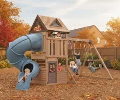 Wooden outdoor playset with a peaked-roof house structure, spiral tube slide, straight slide, climbing wall, and swing set, with several children actively playing in a backyard surrounded by autumn trees, a wooden fence, and a brick house.