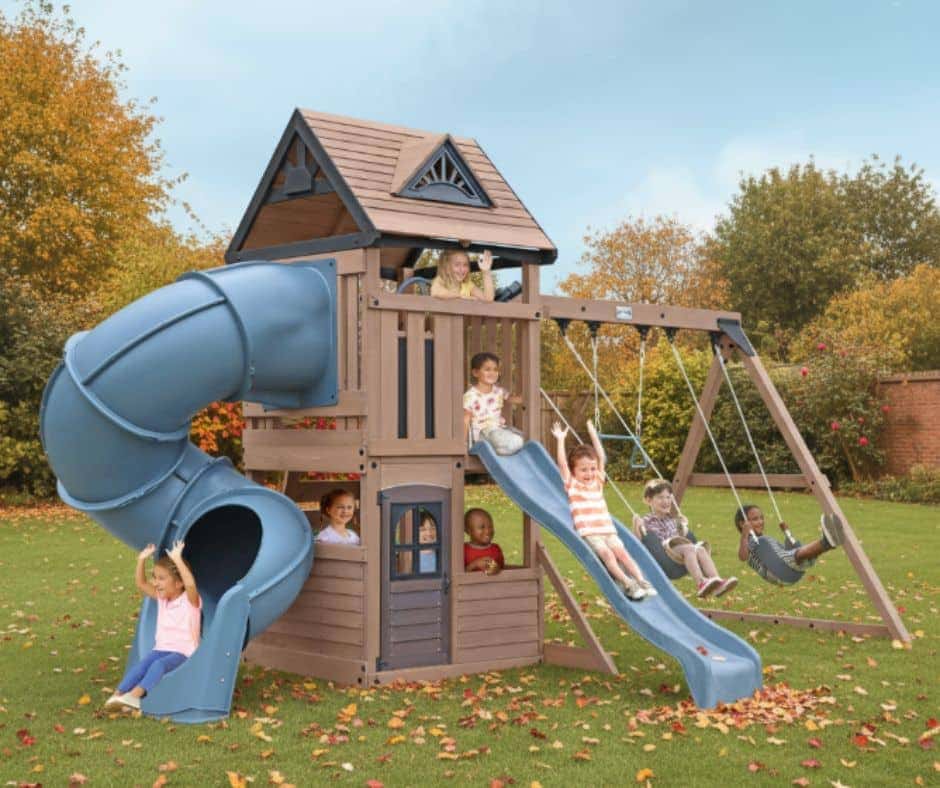 Plastic outdoor playset with a peaked-roof house structure, spiral tube slide, straight slide, climbing wall, and swing set, with several children actively playing in a grassy yard surrounded by autumn trees and a brick wall.