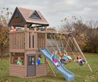 Wooden outdoor playset with a peaked-roof structure, slide, climbing wall, ladder, and swing set, with eight children actively playing in a grassy backyard surrounded by autumn trees in red, orange, and yellow foliage.