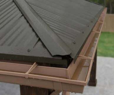 Close-up of a dark corrugated metal roof corner with water droplets and a light brown rain gutter system mounted on wooden beams.