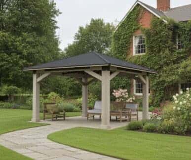 Wooden gazebo with a dark pitched roof supported by beige columns, featuring cushioned wooden benches and chairs arranged on a stone patio, surrounded by neatly trimmed grass, blooming flower beds, and a brick house with ivy-covered walls in the background.