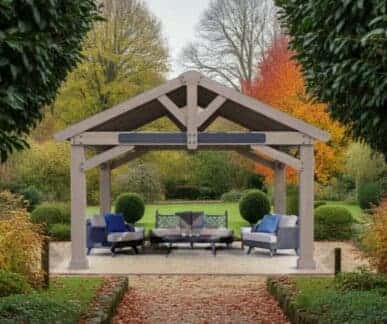 Wooden pergola with a gabled roof and exposed beams, featuring cushioned chairs and a central coffee table, set in a landscaped garden with autumn foliage, manicured hedges, and a stone-bordered pathway.