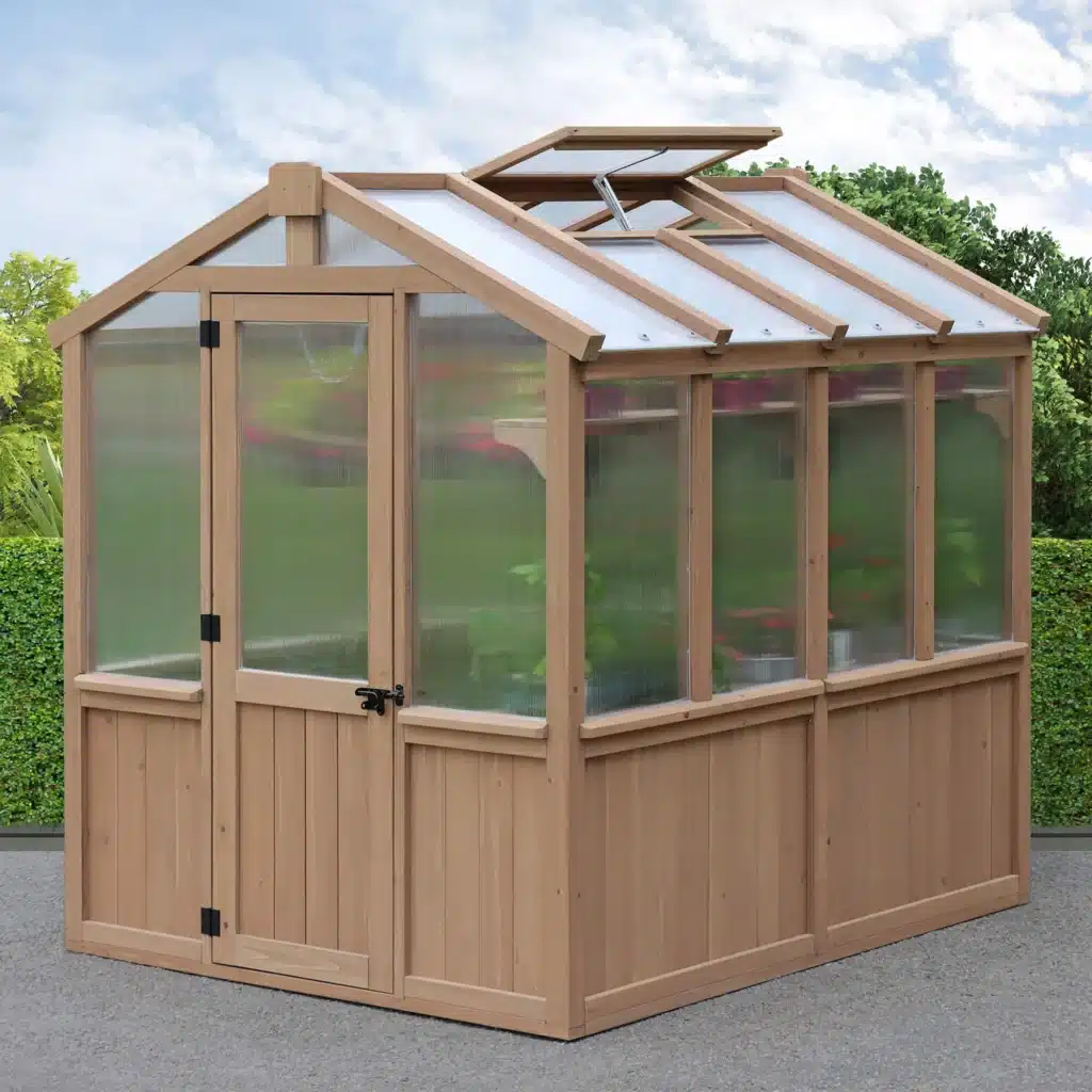 This image features a charming, small-scale wooden greenhouse situated in an outdoor setting, likely a backyard or garden area.
The structure has a classic pitched roof design, similar to a small house or shed. The frame is constructed from light-toned, natural wood, giving it an attractive, rustic appearance. The bottom half of the walls consists of solid wood paneling, while the upper sections and the roof are made with transparent or translucent panels, likely polycarbonate or glass, to allow sunlight in.
The front face of the greenhouse includes a single, wood-framed door with a black handle and latch. The roof has a vent window that is propped open, suggesting ventilation is important for the plants inside. Through the transparent panels, a few green plants can be faintly seen growing inside.
The greenhouse is placed on a level, paved or gravel surface. In the immediate background, a neatly trimmed, vibrant green hedge forms a strong, continuous border. Beyond the hedge, taller trees and bushes with darker green foliage are visible under a bright, partly cloudy blue sky, completing the classic garden scene. The overall look is functional and aesthetically pleasing, designed for personal gardening use.