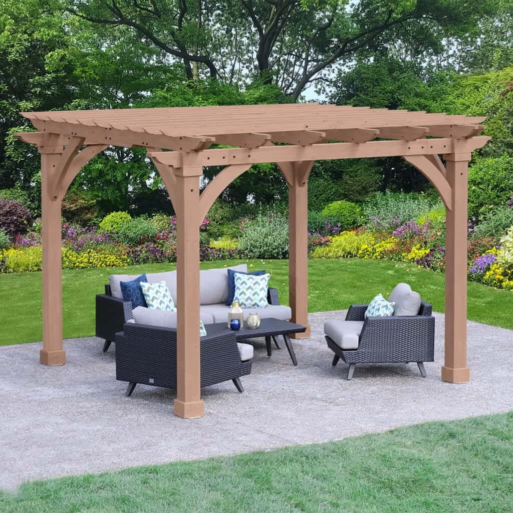 This image showcases a light-toned wooden pergola covering a modern outdoor lounge set on a patio, situated within a lush garden environment.

The pergola structure is made of wood with a warm, natural or light brown stain. It features thick vertical posts and elegant, large arched braces connecting the posts to the horizontal crossbeams, providing a sturdy and aesthetically pleasing design. The roof consists of evenly spaced wooden slats, offering partial shade to the area below.

Underneath the pergola, a contemporary outdoor furniture set is arranged. It includes a sofa and two armchairs, all featuring dark gray or black woven rattan frames and thick, light gray cushions. The set is centered around a rectangular dark coffee table. The furniture is accented with decorative throw pillows in shades of white, light blue, and green, adding pops of color and pattern.

The furniture rests on a level patio of light gray concrete or paving stones. This patio is surrounded by a vibrant green lawn in the foreground. The background features a beautifully maintained garden bed overflowing with colorful flowering shrubs and perennials in shades of pink, yellow, and purple. Tall, mature trees provide a deep green backdrop against the brightly lit scene, emphasising the pergola as a perfect focal point for relaxation and entertaining in the garden.