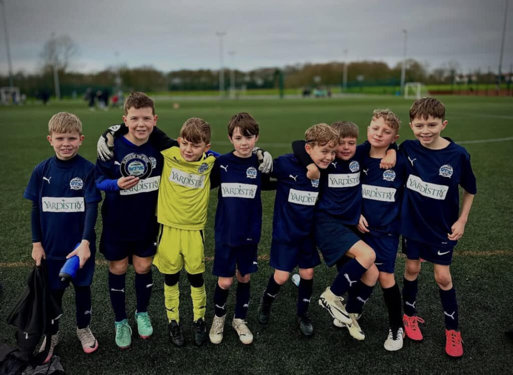 This image captures a group of nine young boys posing together on an artificial turf soccer field after what appears to be a match or tournament.

The boys are dressed in soccer uniforms. Most of the team is wearing dark blue shirts and shorts with the word "YARDISTRY" prominently featured as a sponsor name in white or light yellow text across the chest. The boys are also wearing matching dark socks and various types of soccer cleats. One player, positioned slightly to the left of the center, is wearing a bright yellow goalkeeper kit (jersey, shorts, and socks) that also displays the same sponsor logo.

The boys are standing close together in a line, many with their arms around each other's shoulders, conveying a sense of camaraderie and team spirit. Their expressions are cheerful, indicating happiness or pride. The second boy from the left is holding a small, dark blue trophy or plate, suggesting they may have won an award or reached a milestone. In the center-right, two boys are engaged in a playful, supportive pose, with one boy being held slightly off the ground by his teammate.

The setting is an outdoor, well-maintained synthetic turf sports field under an overcast sky, suggesting cool or cloudy weather. In the background, beyond the playing area, there is a clear boundary, and in the far distance, some trees and buildings are softly visible. The overall impression is a candid, joyful team photograph.