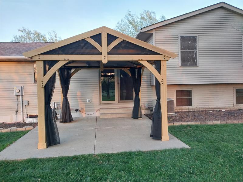 Wooden backyard gazebo with mesh curtains on concrete patio beside beige two-story house.